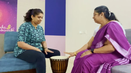 Two women sit opposite each other playing a drum